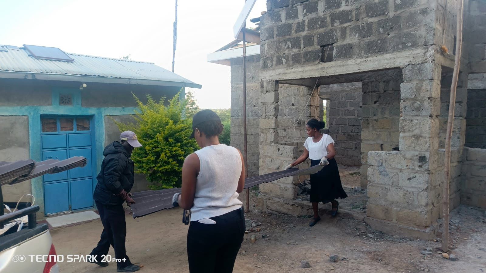Customer and crew carrying brown tile-profile mabati sheets into a stone house under construction