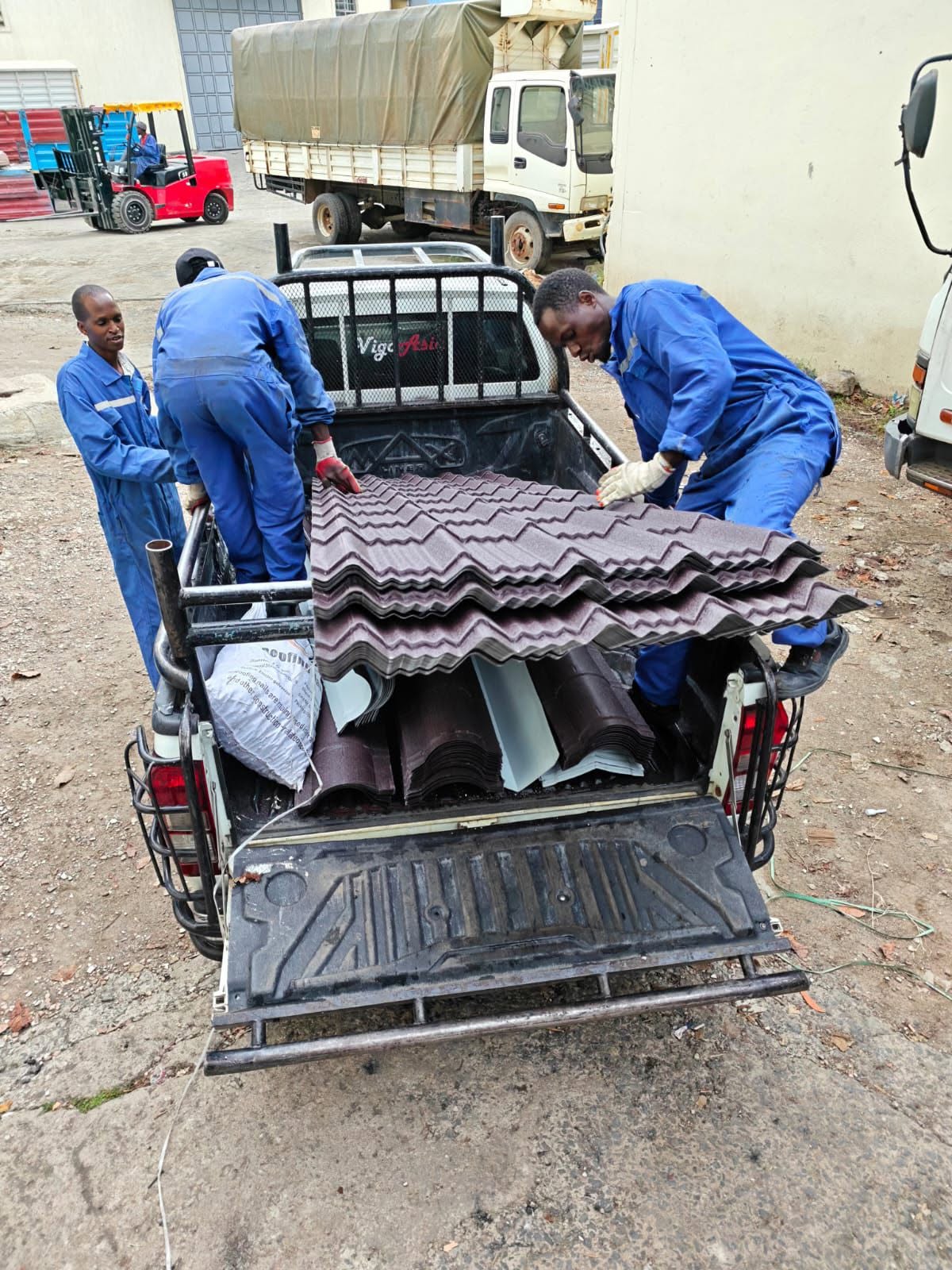 Three Tranquility Mabati staff in blue overalls loading maroon stone-coated tiles onto a customer pickup